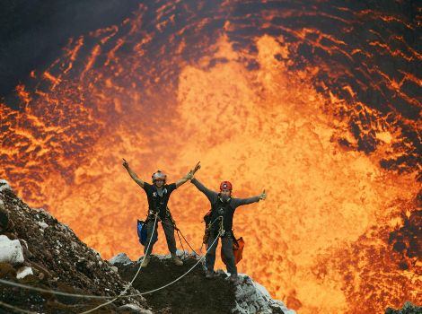Two men are suspended on ropes above an active volcano crater.