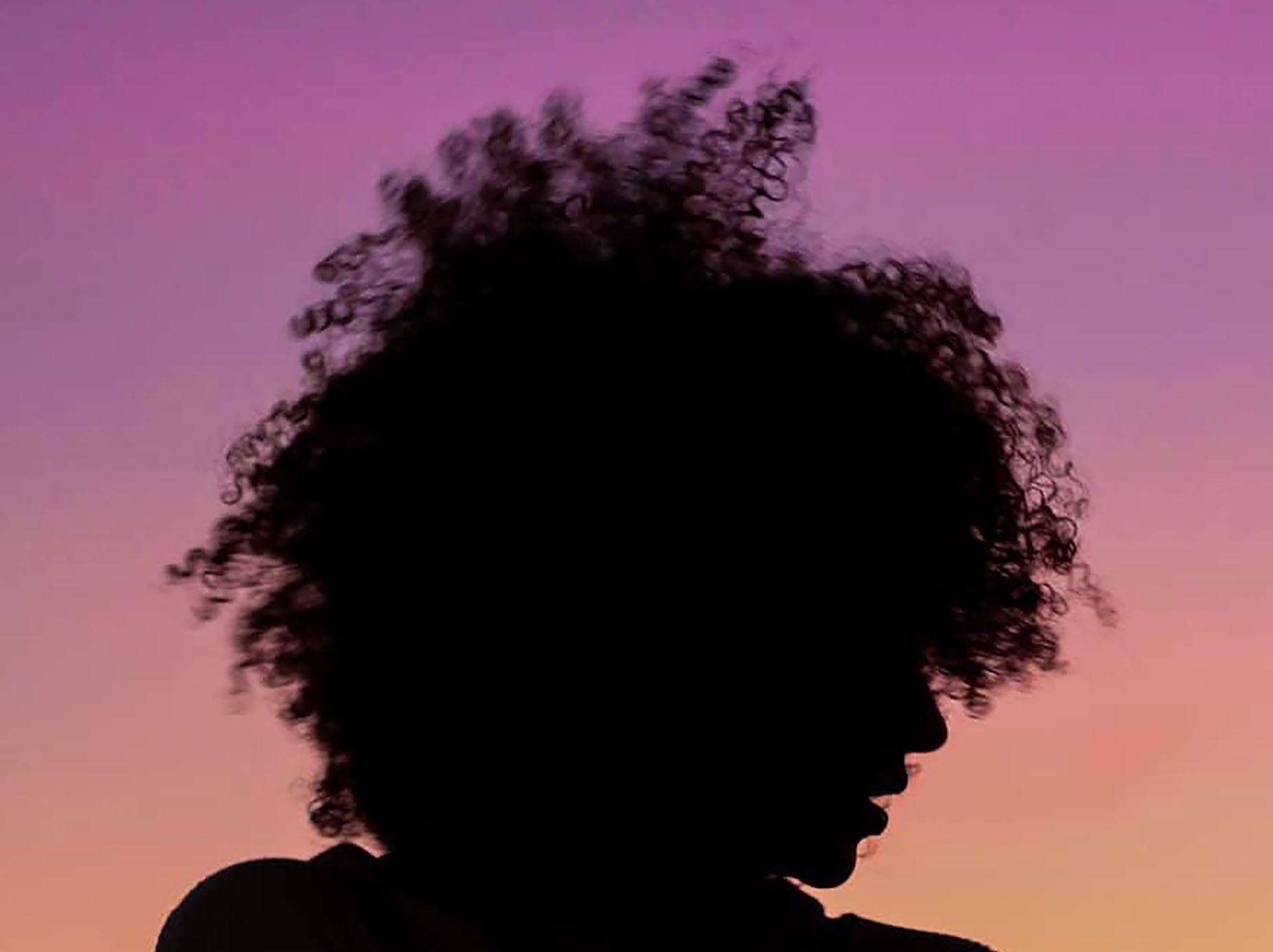 A woman's face, silhouetted against the sky, her curly hair outlined by red and purple hues of the sunset.