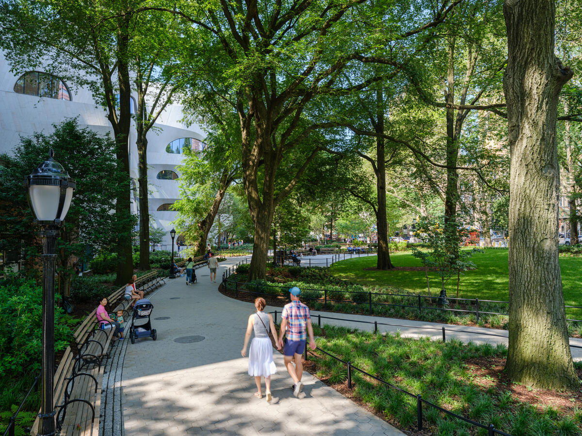 Two visitors holding hands, one in a plaid shirt and another in a white skirt, walking down a path facing the Gilder Center in the Theodore Roosevelt Park on a bright and sunny day.