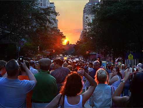 A large crowd dressed in bright clothing looking at a bright yellow and orange sunset with several individuals taking photographs with cameras or phones.