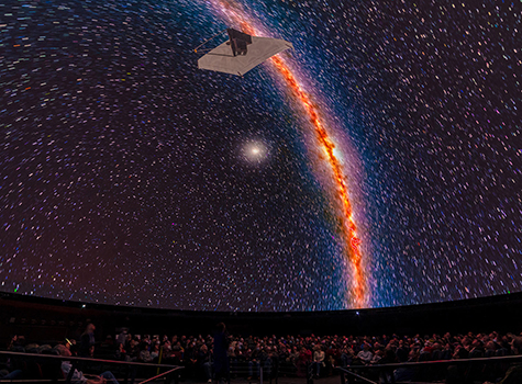 A wide angle photograph of an audience in the Hayden Planetarium, looking up at a depiction of a spacecraft against white stars. 