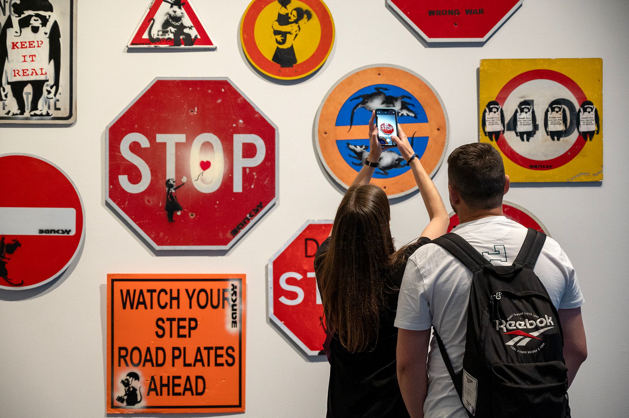 Two people stand in front of a wall of Banksy artwork, which includes a Stop sign, circular signs, and a sign reading 'WATCH YOUR STEP ROAD PLATES AHEAD'