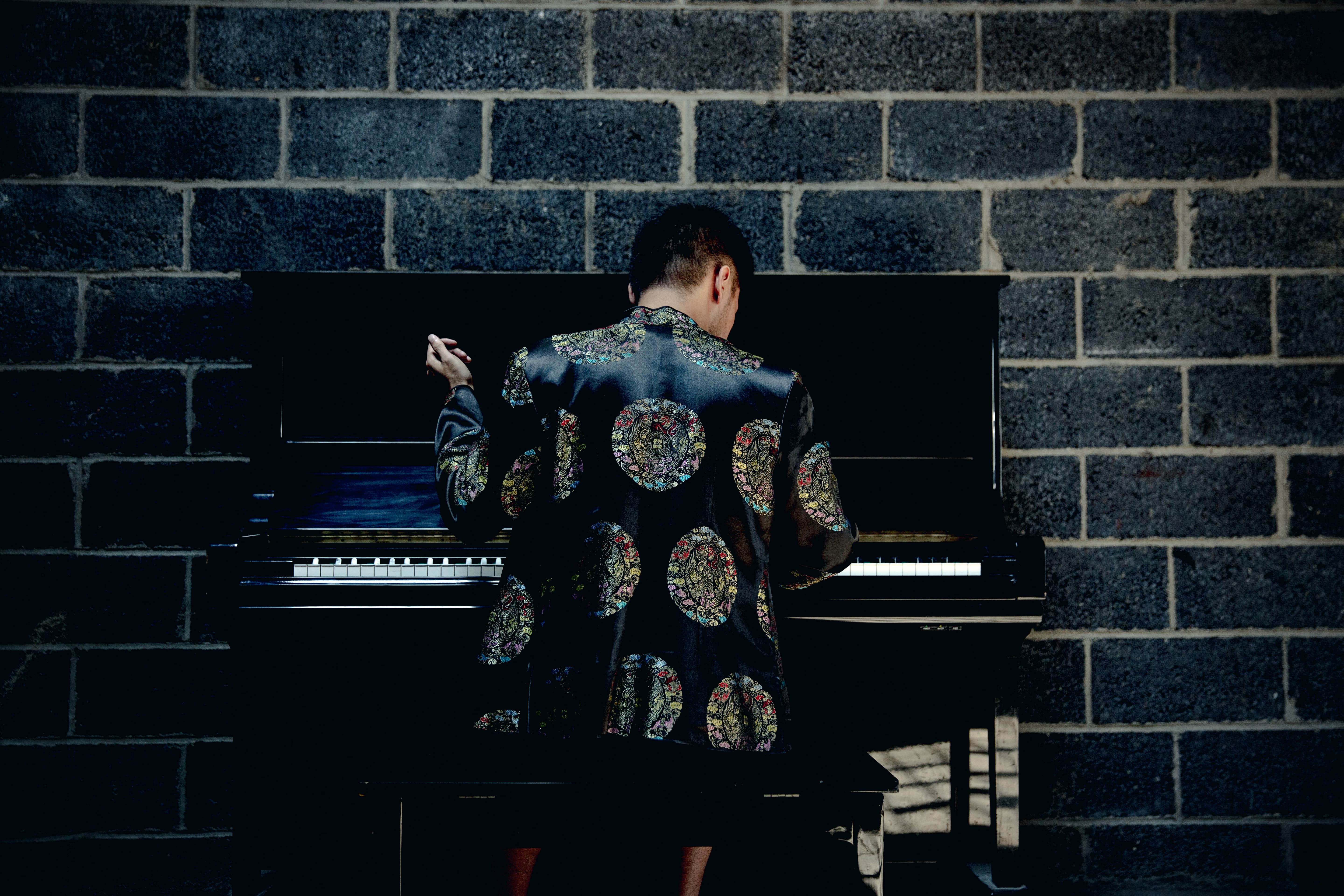 We view Conrad Tao from the back. He is wearing an embroidered jacket and seating at a piano.