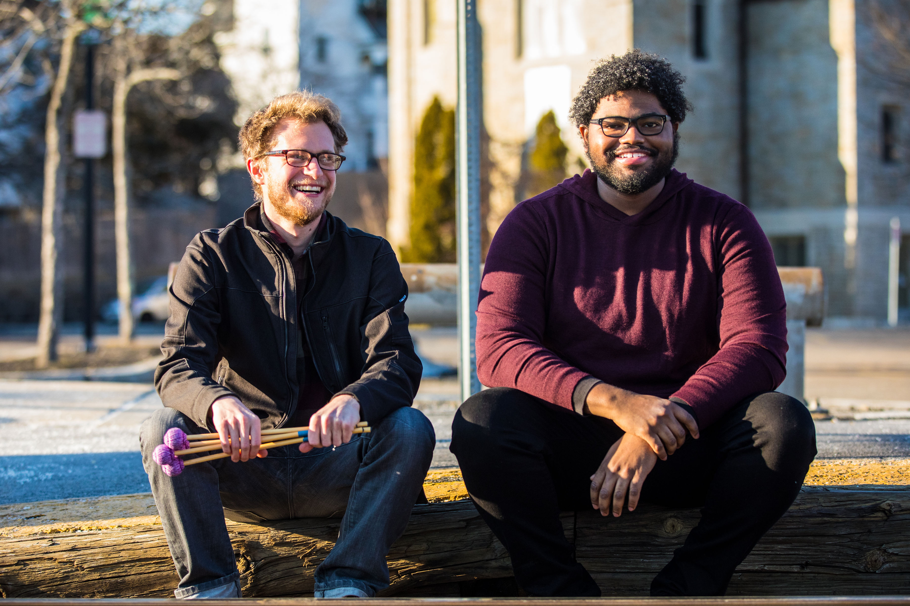 ID: Julian Loida and Charles Overton are sitting on a curb. Loida holds his mallets in his hands and looks to his left. Overton rests his hand on his arm as he looks forward.