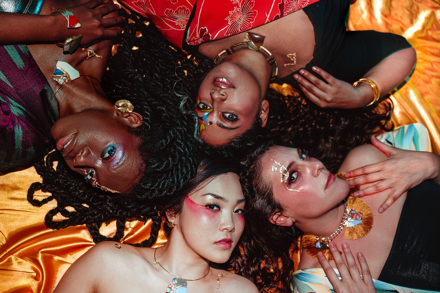 ID: Débo Ray, Annette Philip, Giorgia Renosto, and Ayumi Ueda are laying clockwise over an orange satin sheet. Their brown hair blends together in the center.