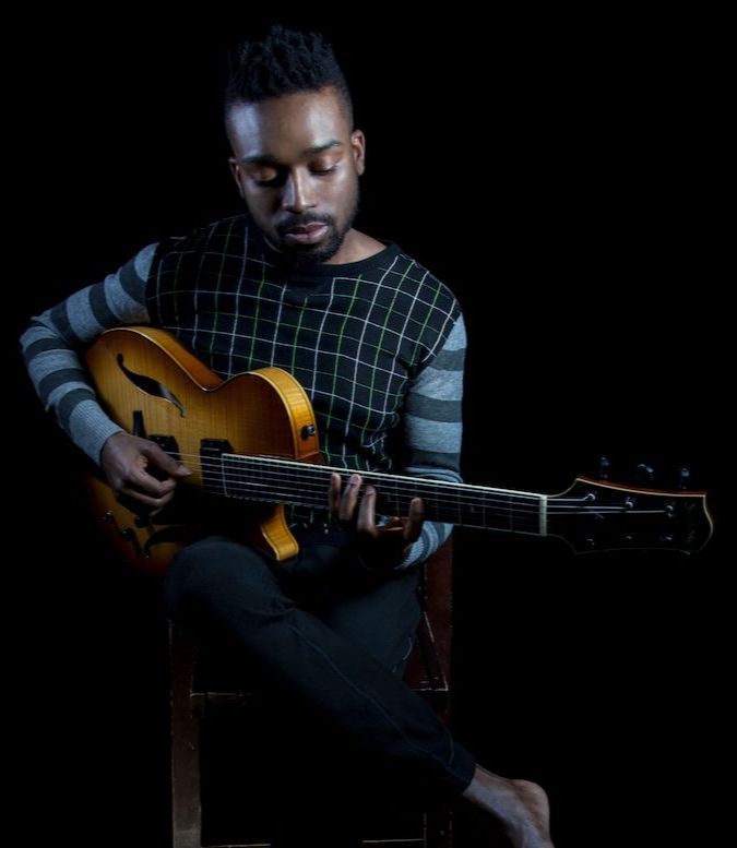 Albino Mbie is sitting on a stool, playing an electric guitar