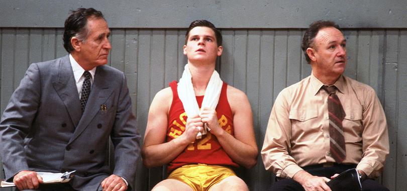 A still from the movie Hoosiers with a coach in a grey suit and basketball players in gold jackets.