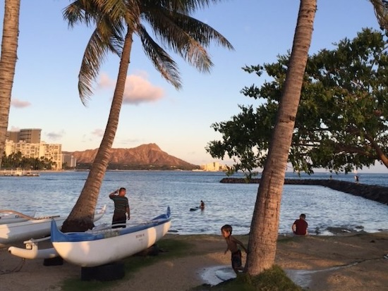 A photo of a Hawaiian beach scene, with a few people and a boat on the beach beside palm trees and high-rise buildings across the water