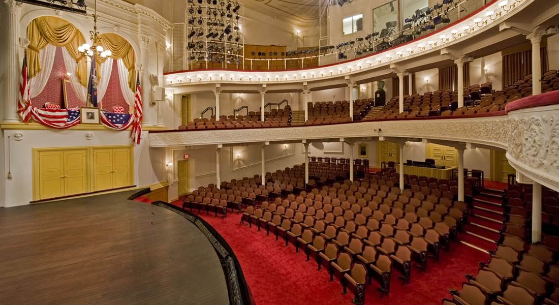 A side view of the stage and seating at Ford's Theatre. On the left is the President's Box with an American flag, a framed picture of George Washington and American flag bunting draped over the box. Link to Welcome Back information.