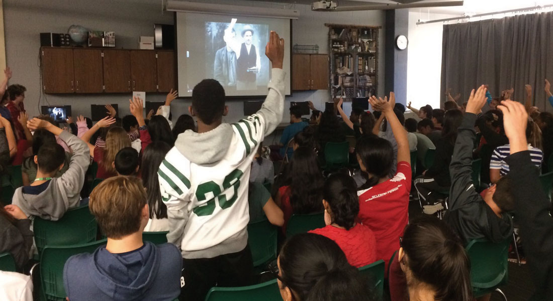 A large classroom full of students watching an actor portraying Detective McDevitt on a projector screen. Many students are raising their hands to participate. 