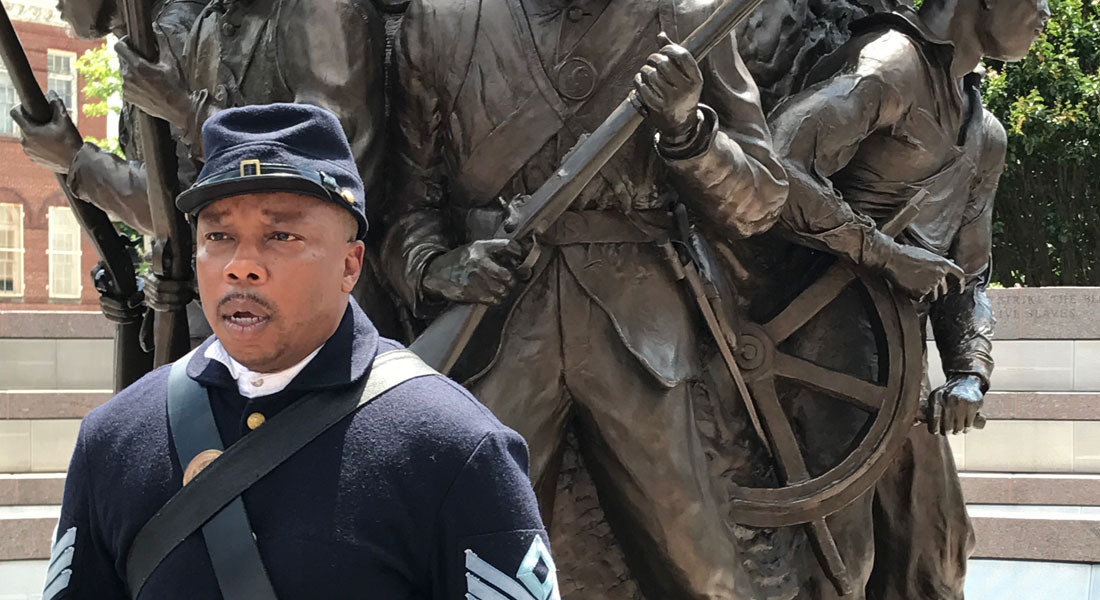 An African American man, dressed in a Union Uniform, standing in front of a bronze military memorial sculpture depicting four different African American men from each branch of the military. 