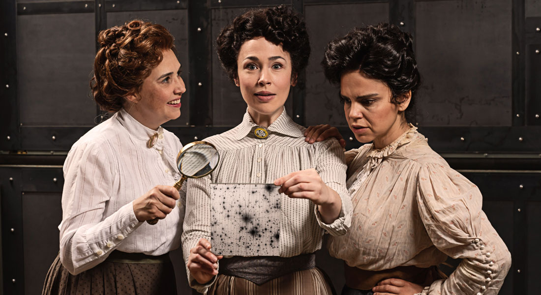 Three actresses wearing Victorian blouses and high-waisted skirts stand together. The woman in the middle holds a glass plate with a photograph of the night sky and has an expression of discovery on her face. Link to synopsis.