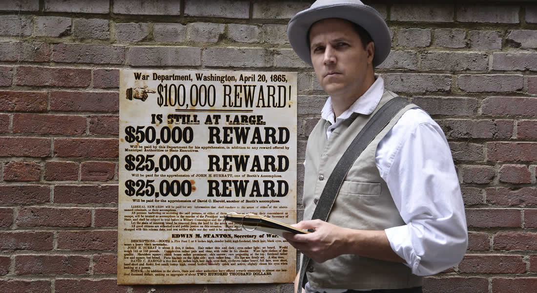 A man, dressed in 1860s costume and holding a detective's notebook, stands in front of a brick wall and Wanted Poster for John Wilkes Booth. 