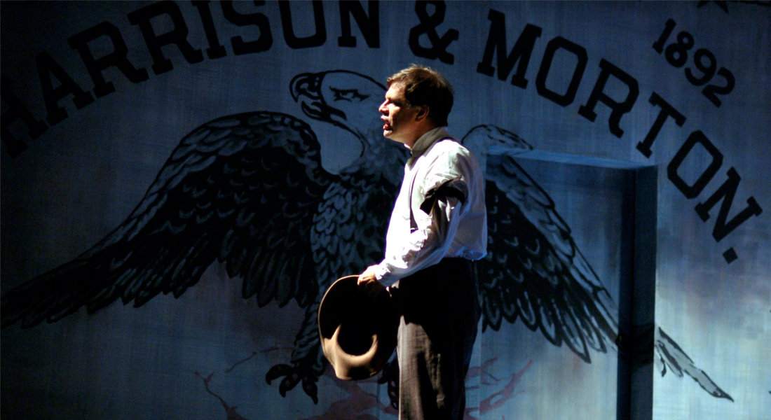 Profile photo of a man standing while holding his hat in front of a dramitcally lit mural with an eagle with it's wings spread. Link to gallery.