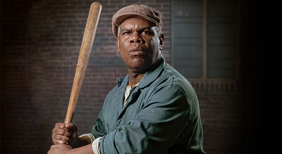 Actor playing August Wilsons character Troy from the play Fences stands in his 1950s costume. Troy is holding a baseball bat as if he were about to swing while wearing faded-green overalls and a tan linen cap.