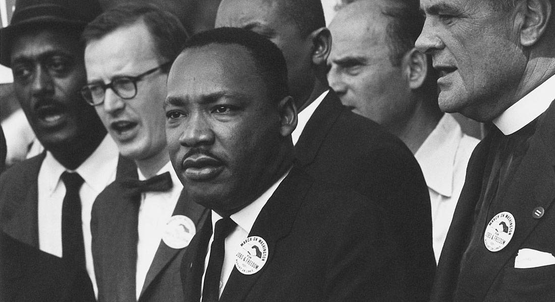 A black-and-white photo of Martin Luther King Jr. He wears a black suit, white shirt and black tie. He stands next to and in front of men carrying protest signs. Link to upcoming season.