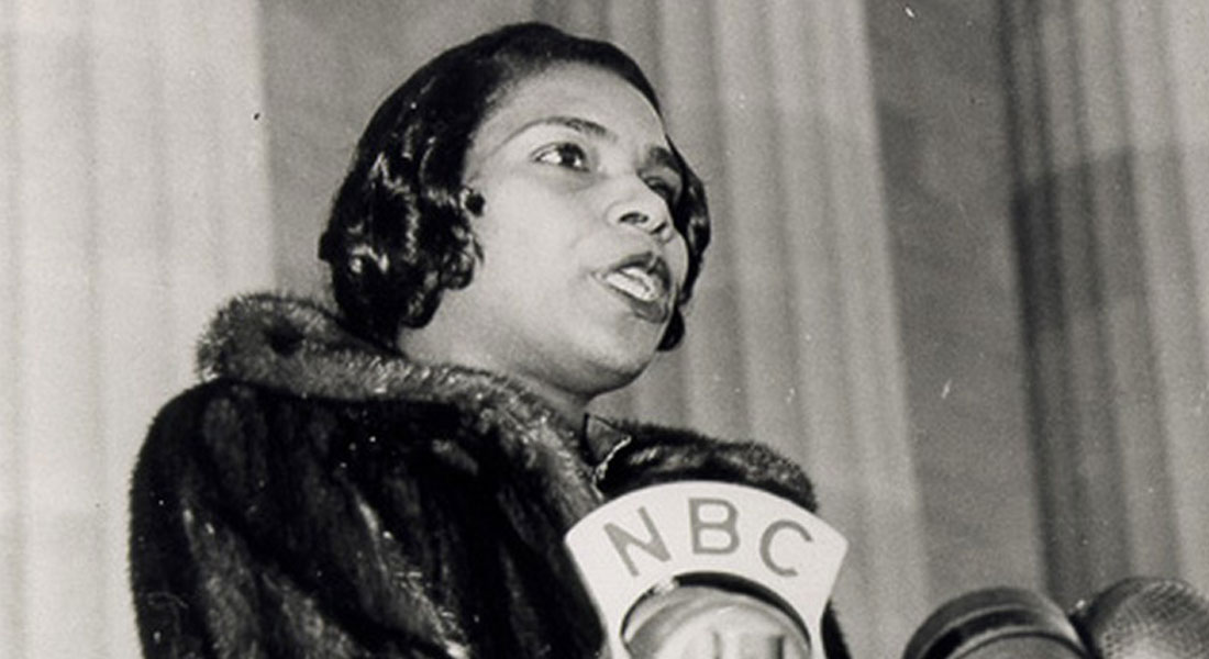 A black-and-white photo of Marian Anderson. She wears a fur coat and sings into several large microphones while standing in front of the columns of the Lincoln Memorial. Link to upcoming season.