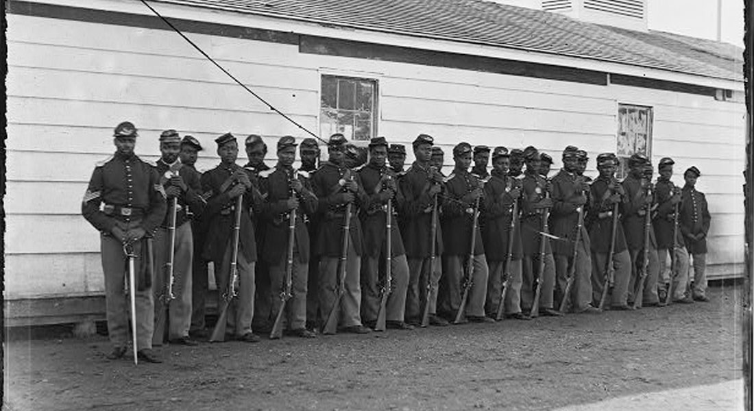 26 African American Civil War soldiers in uniform, pose in line in front of a barracks.