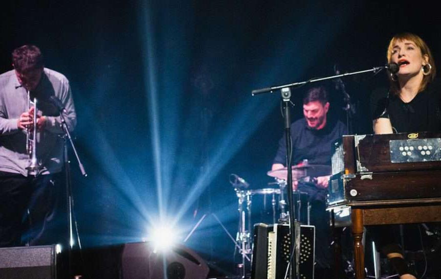 Three people are on stage performing. A young woman is to the right of the image singing into a microphone and playing a piano. A young man with brown hair is behind her playing drums, and a young man to the left of the image is playing trumpet