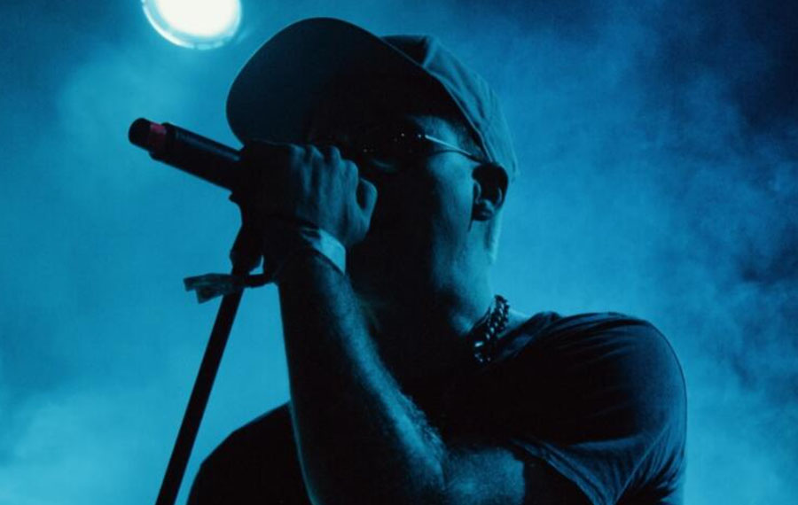A young man with dark hair and a cap is speaking into a microphone. He is wearing a grey t shirt and there is blue smoke behind him in the background