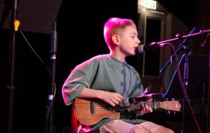 A young boy with short blonde hair is sat down playing a ukulele and singing into a microphone. He is wearing a green t shirt