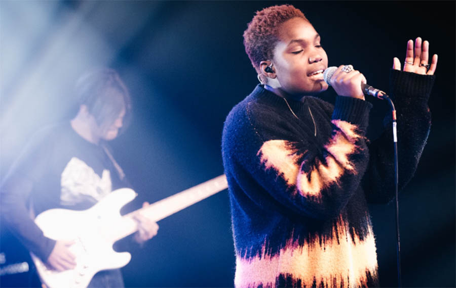 Two individuals are seen performing on stage. One is a young woman with short brown hair playing drums and the other is a young man with dark hair playing a keyboard