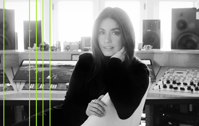 A young woman with medium-length brown hair is sat down with her chin resting on her arm. She is looking straight into the camera. There is a music studio desk behind her