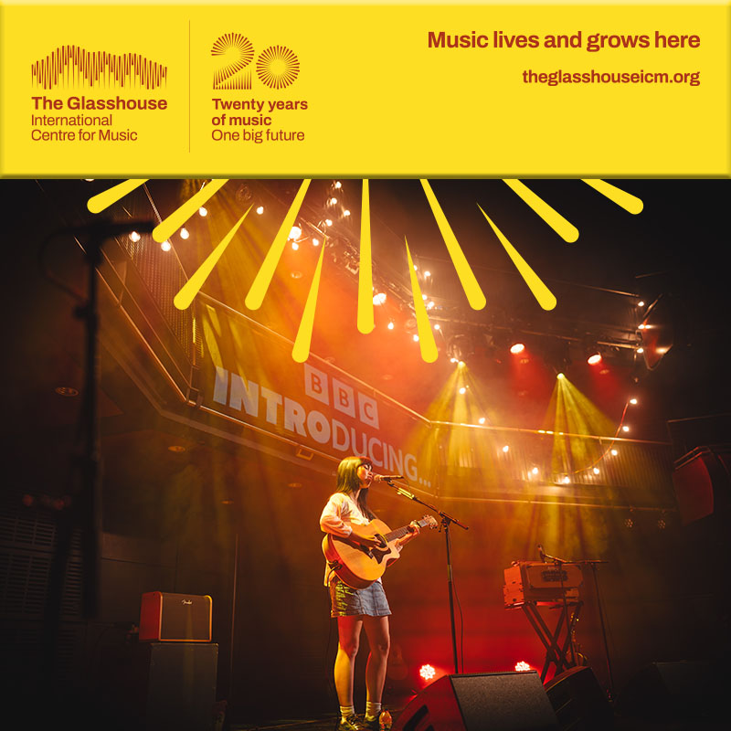 Nadia Kadek: A young woman wearing a denim skirt and long sleeve top singing and playing an acoustic guitar on stage.