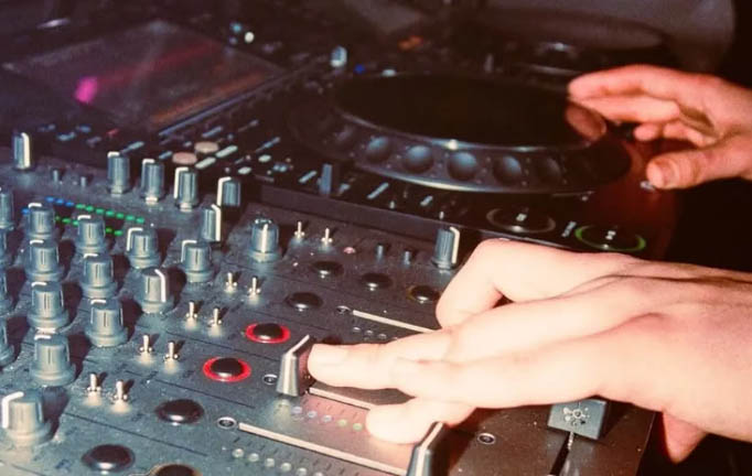 Two hands are seen - one pushing up a slider on a mixing desk and the other on a DJ booth. 