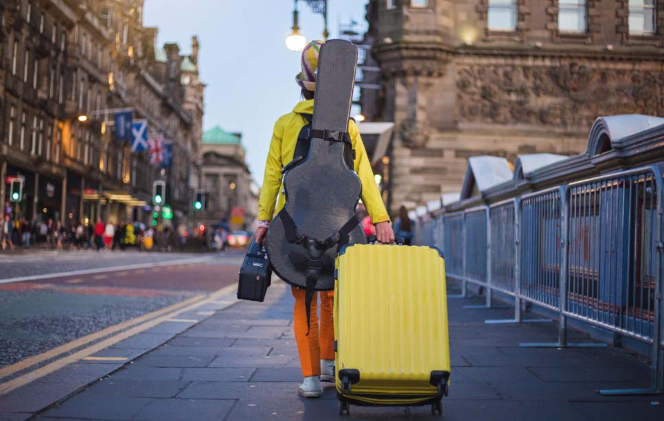 An individual is seen walking away from the camera, carrying a guitar on their back and pulling a yellow suitcase in their right hand