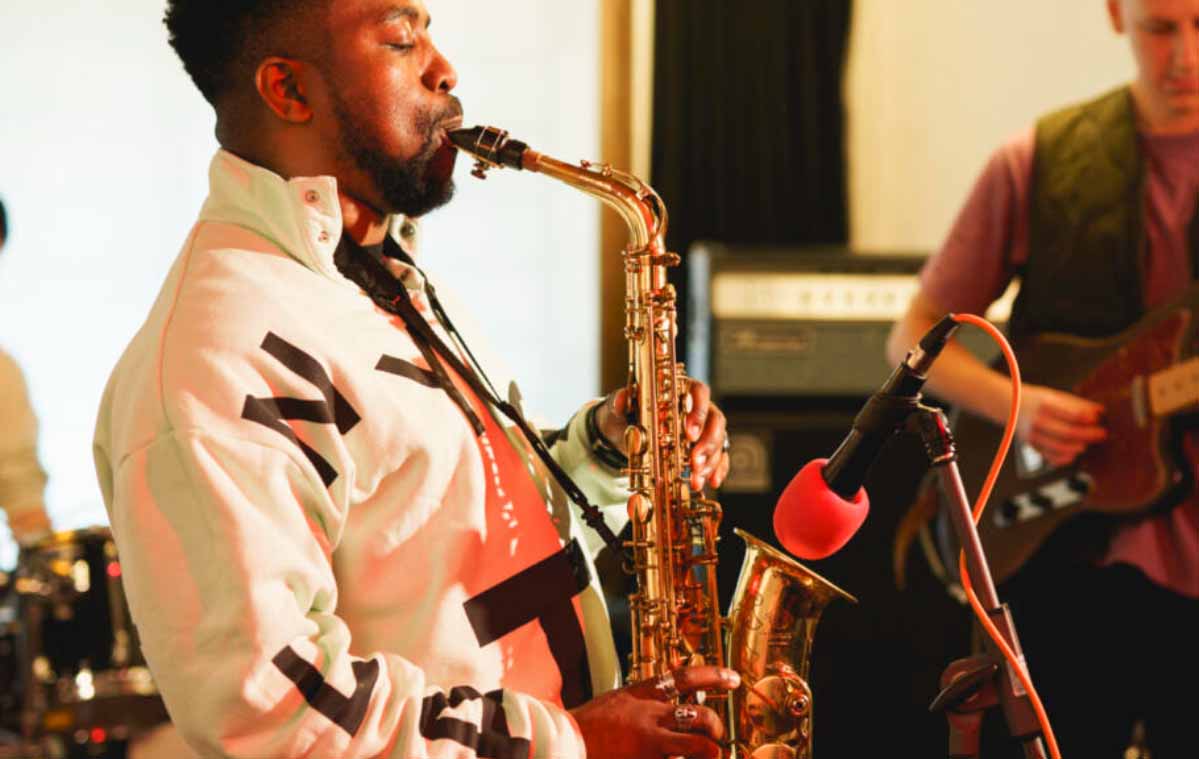 A young man is seen standing and playing saxophone. The man has dark hair and a white jacket on
