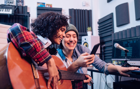 Two young women are sat down together laughing. They are both looking at a mobile phone which the person on the right is holding. She is wearing a beanie hat and a blue long sleeved shirt. The other woman is holding an acoustic guitar. She has brown hair.