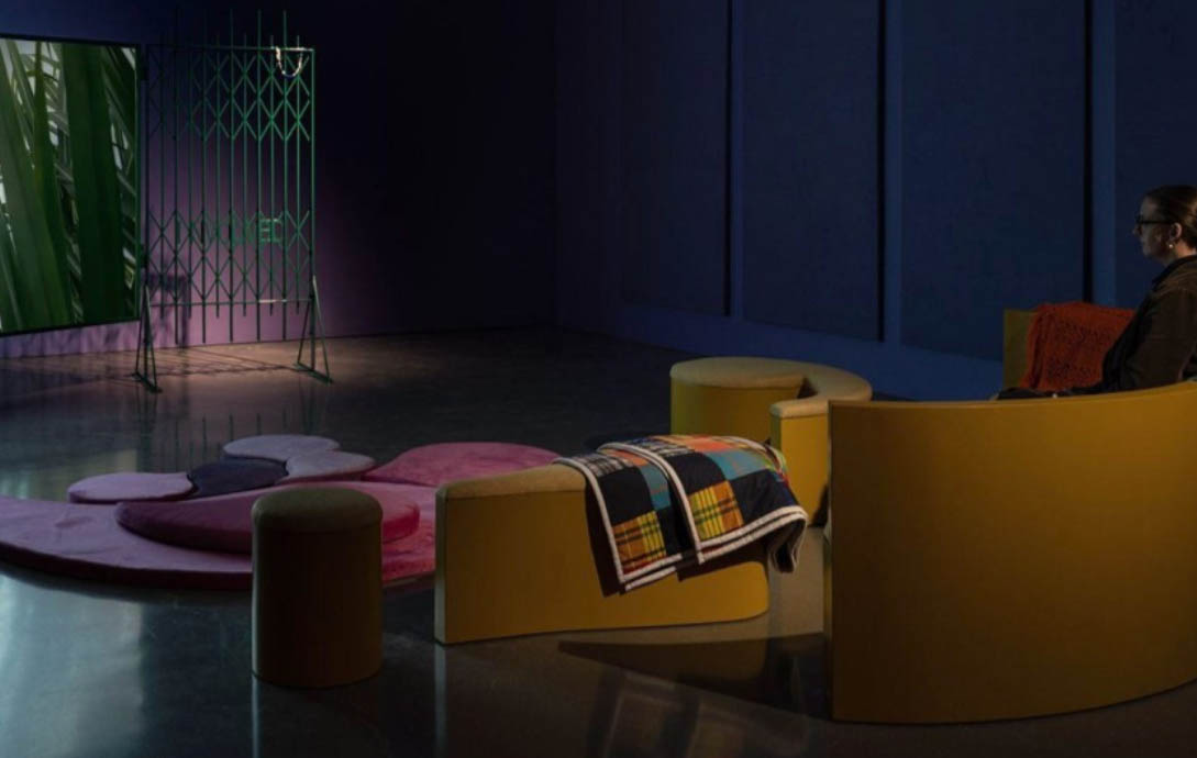 A line of mustard colour seats are shown at the front of the image. There is a patterned blanket draped over the centre seat. In the top right hand corner, there is a TV screen with an image of plants. A pink and purple rug is on the floor in front of the seats.