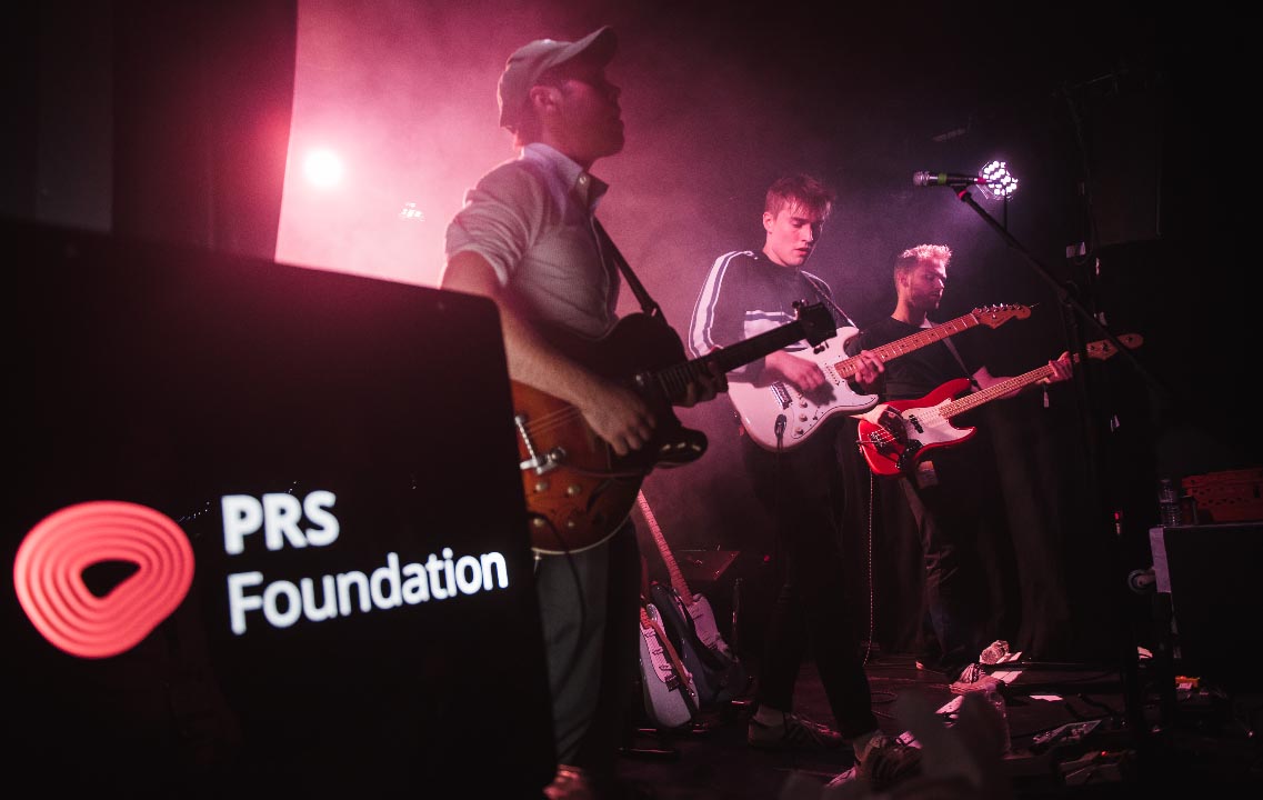 Three young men are stood centre stage, performing and playing electric guitars. The man in the middle is Sam Fender. The man closest to the camera is wearing a grey cap and a long sleeved top with his sleeves rolled up. The PRS Foundation logo is to the left of the image.
