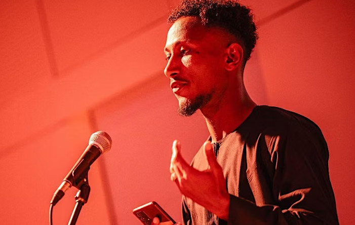 A young man is standing in the centre of the image. He is speaking into a microphone. He has dark hair and a beard. He is holding a phone in his right hand and holding his left hand up - making a gesture as he explains. He is wearing a brown long sleeved top.