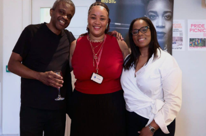 Three individuals are stood smiling at the camera. They are linking arms with each other. 