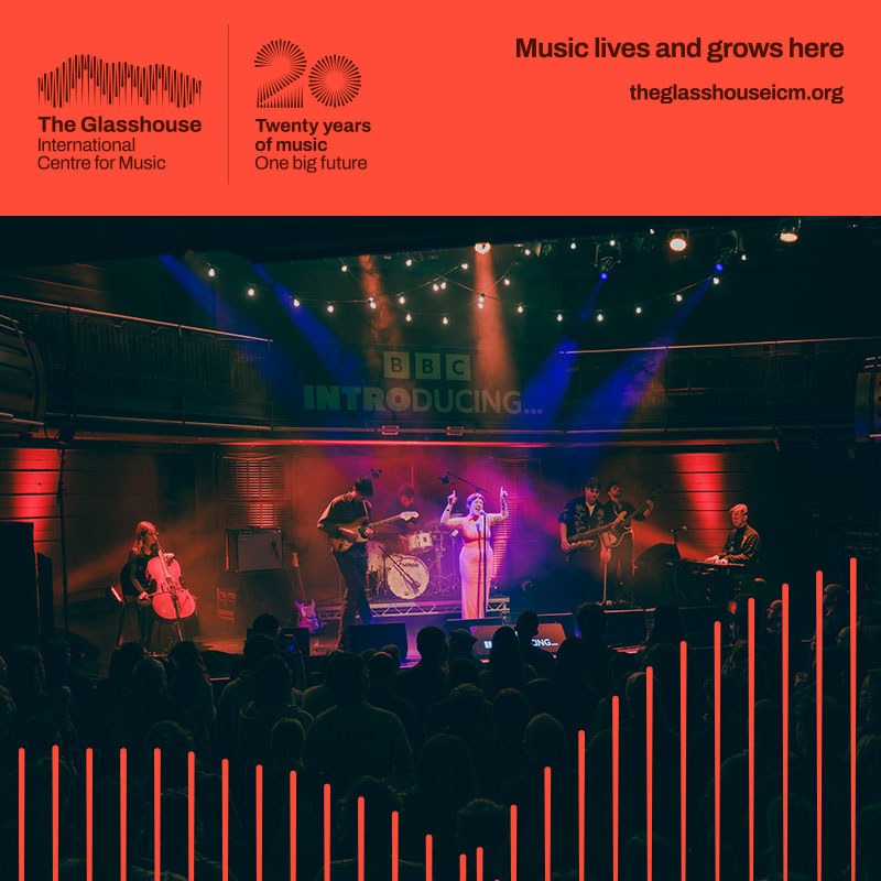 Ernie: a band of six people are performing on stage with red lighting behind them. Three are singing into microphones, two are playing electric guitars, and one is playing drums at the back