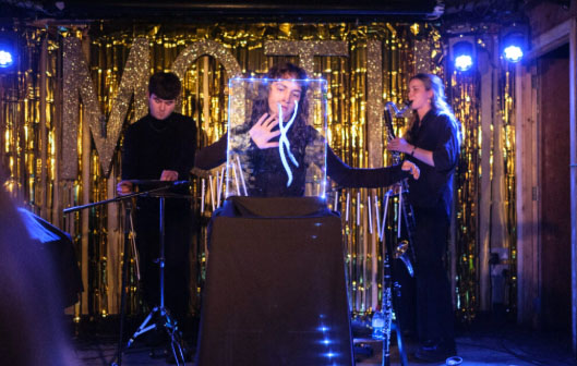 One girl is stood to the right of the image playing a saxophone. Another girl is stood in the centre behind a small screen. A boy is stood to the right of the image playing a MIDI keyboard. They are all wearing black outfits