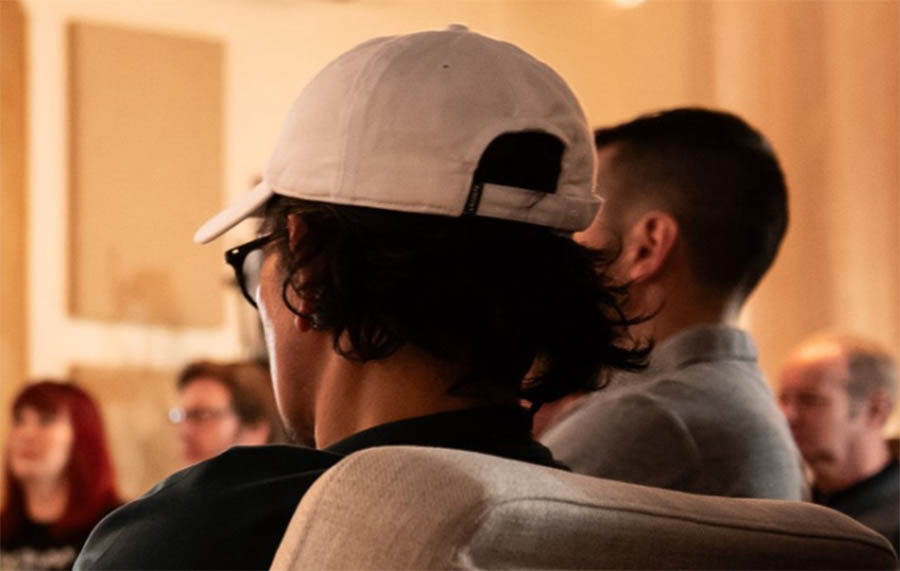 A young man with black curly hair is sat facing away from the camera. He is wearing a grey cap and black glasses. There are also other people out of focus in the background of the image