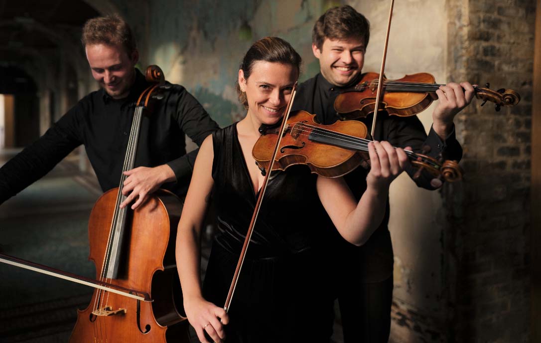 Two people are standing playing violins. They are smiling directly into the camera. Another man stands behind them playing a cello