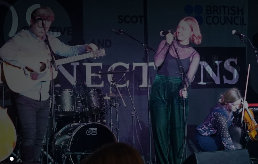 Three individuals are seen on stage. The person to the left of the image is tuning their acoustic guitar and wearing a long sleeved blue shirt with dark jeans. The person centre stage is holding the microphone stand and wearing green trousers and a black mesh top. The person to the right of the image is crouched down holding a violin and bow