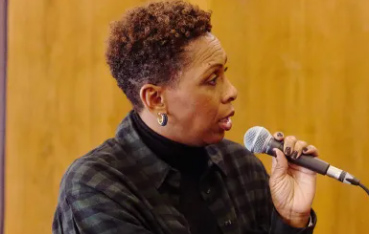 A woman is sat down looking to her left as if speaking to someone. She is holding a microphone and is wearing a green checked shirt with green earrings