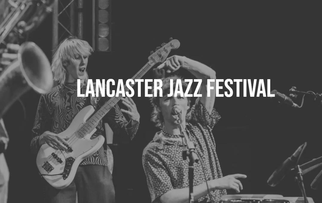 A young man is sat down, singing into a microphone and playing a drum. Another person is stood behind him playing the bass guitar. The wording 'Lancaster Jazz Festival' is written over the top of this image