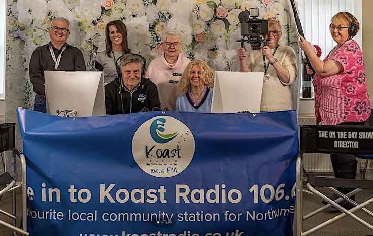 A group of people are sat behind a banner smiling into the camera. One of the women is holding a video camera and another woman has a boom mic for audio