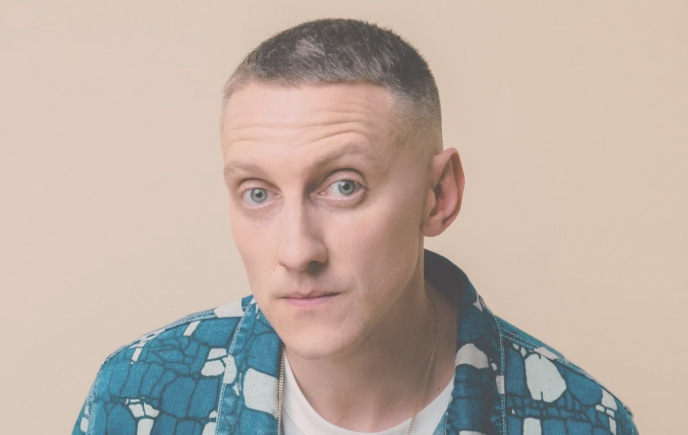 A young man is staring directly into the camera. He is wearing a blue and white jacket and has very short hair