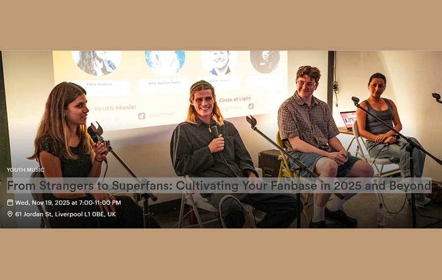 Four individuals are seen speaking into microphones and sitting on chairs looking out into the audience. Two are female and two are male. They are all smiling.