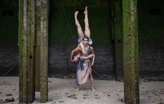One woman is stood in the middle of the image looking directly at the camera. She is lifting up a man who is upside down. Sand covers the floor and four beams are surrounding the pair