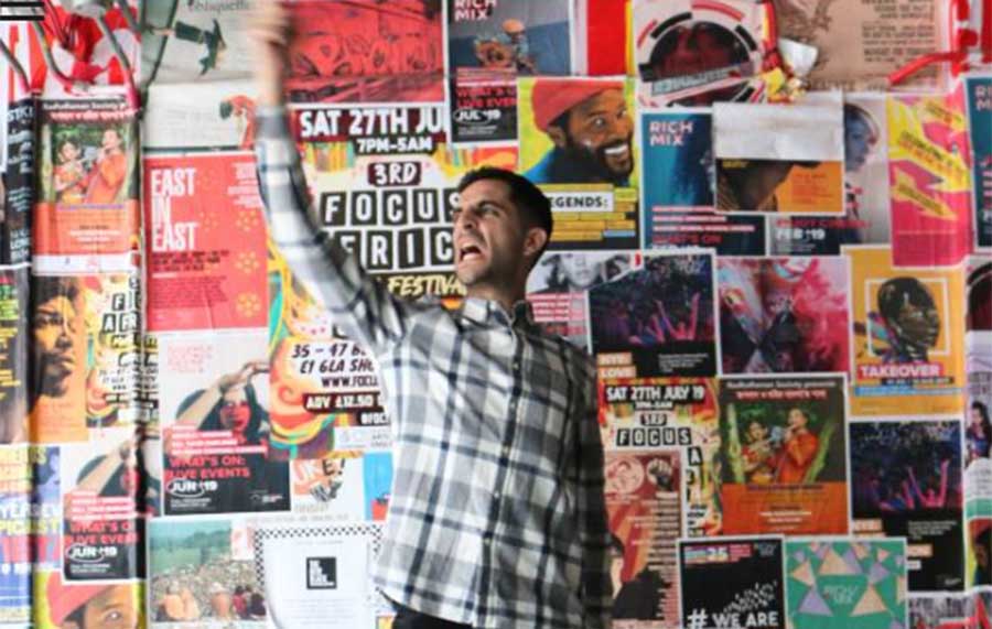 A young man with dark hair wearing a checked shirt is seen punching his fist into the air. The background is full of various magazine covers