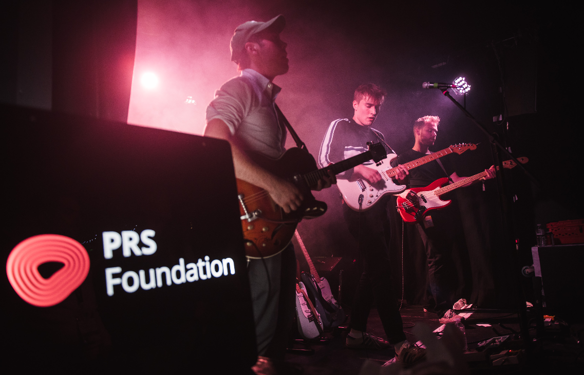 Three young guys are seen playing electric guitars on stage. The PRS Foundation logo is to the front of the image on the left