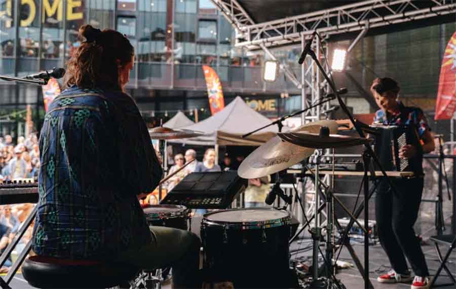 Two individuals are seen performing on stage. One is a young woman with short brown hair playing drums and the other is a young man with dark hair playing a keyboard